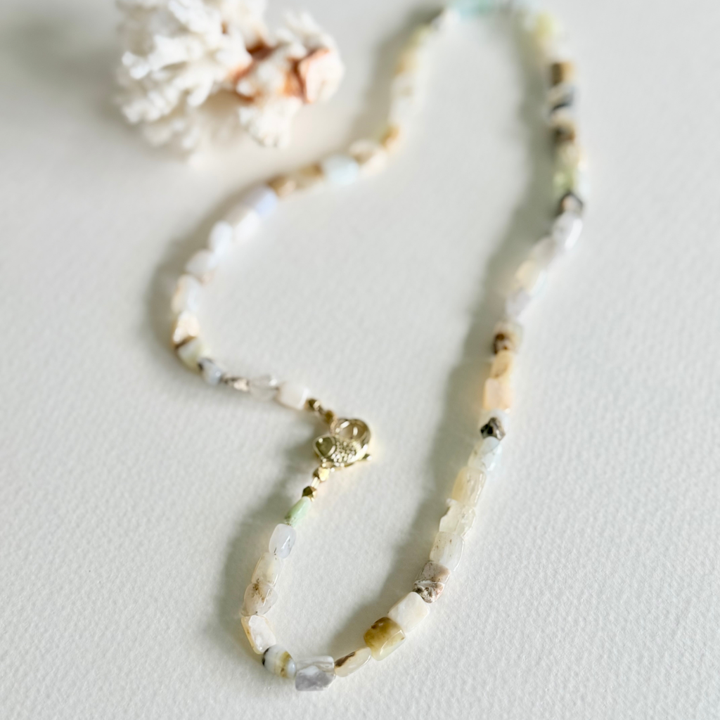 A beaded necklace made of small natural stone beads in soft cream, beige, white, and pale green tones, with a small gold clasp, photographed on a light neutral surface with a decorative coral-like prop near the top left.
