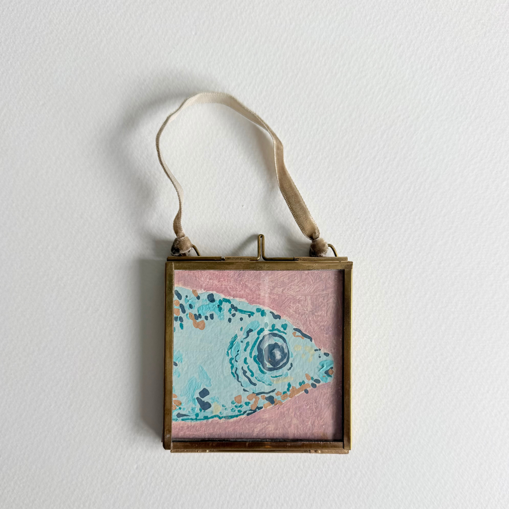 A square framed wall hanging with a canvas painting of a stylized fish in light blue and turquoise on a pink background, enclosed in a brass frame with a beige hanging ribbon, shown against a white wall.