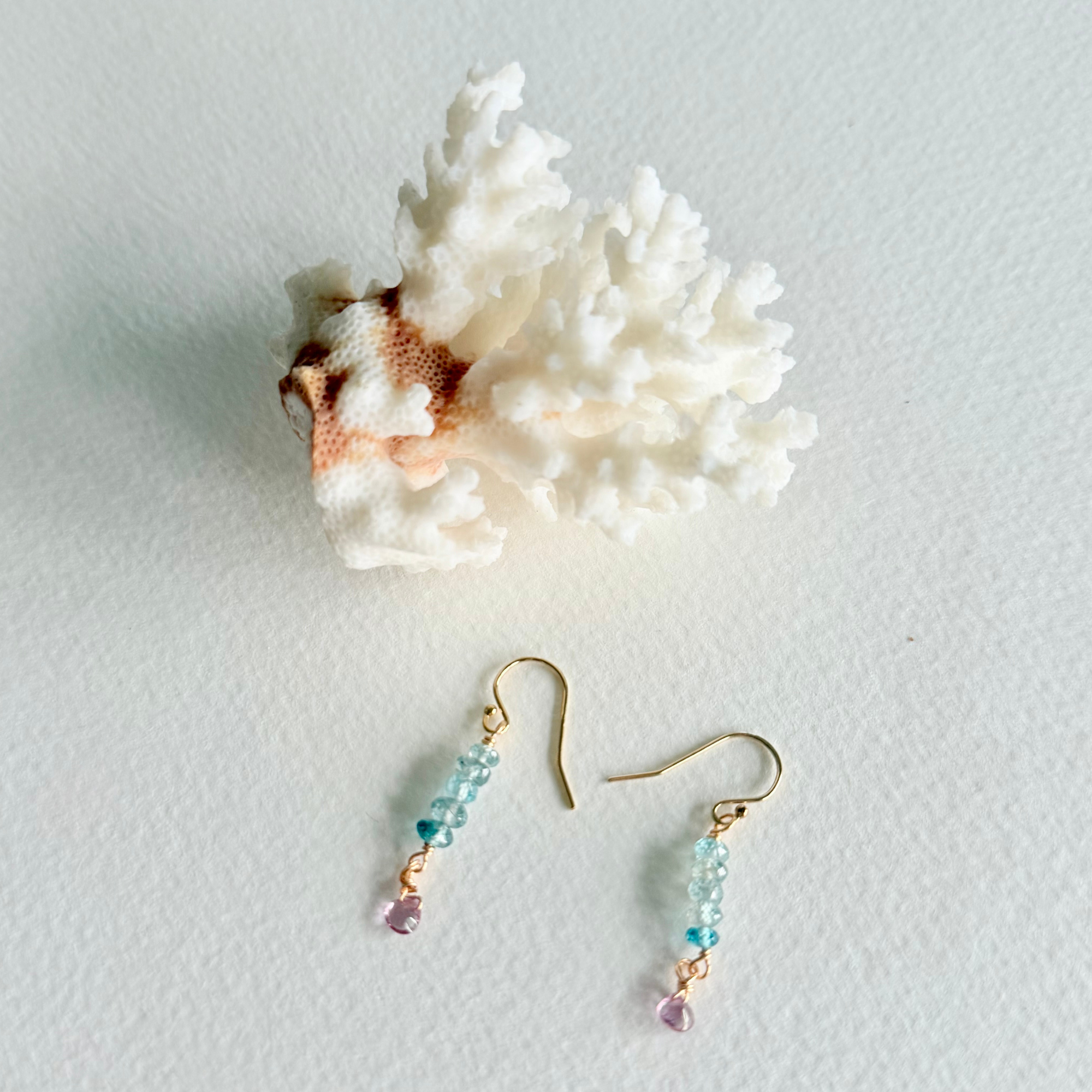 A pair of gold-tone dangle earrings with light blue and purple beads, displayed on a white textured surface next to a white coral shell.