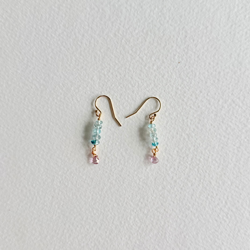 A pair of small dangle earrings with gold-tone hooks, light blue faceted beads, and tiny pink dangling accents, photographed on a plain light background.