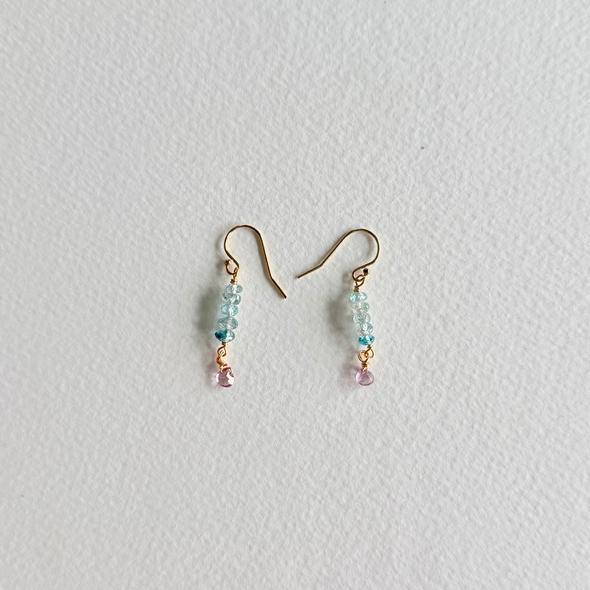 A pair of small dangle earrings with gold-tone hooks, light blue faceted beads, and tiny pink dangling accents, photographed on a plain light background.