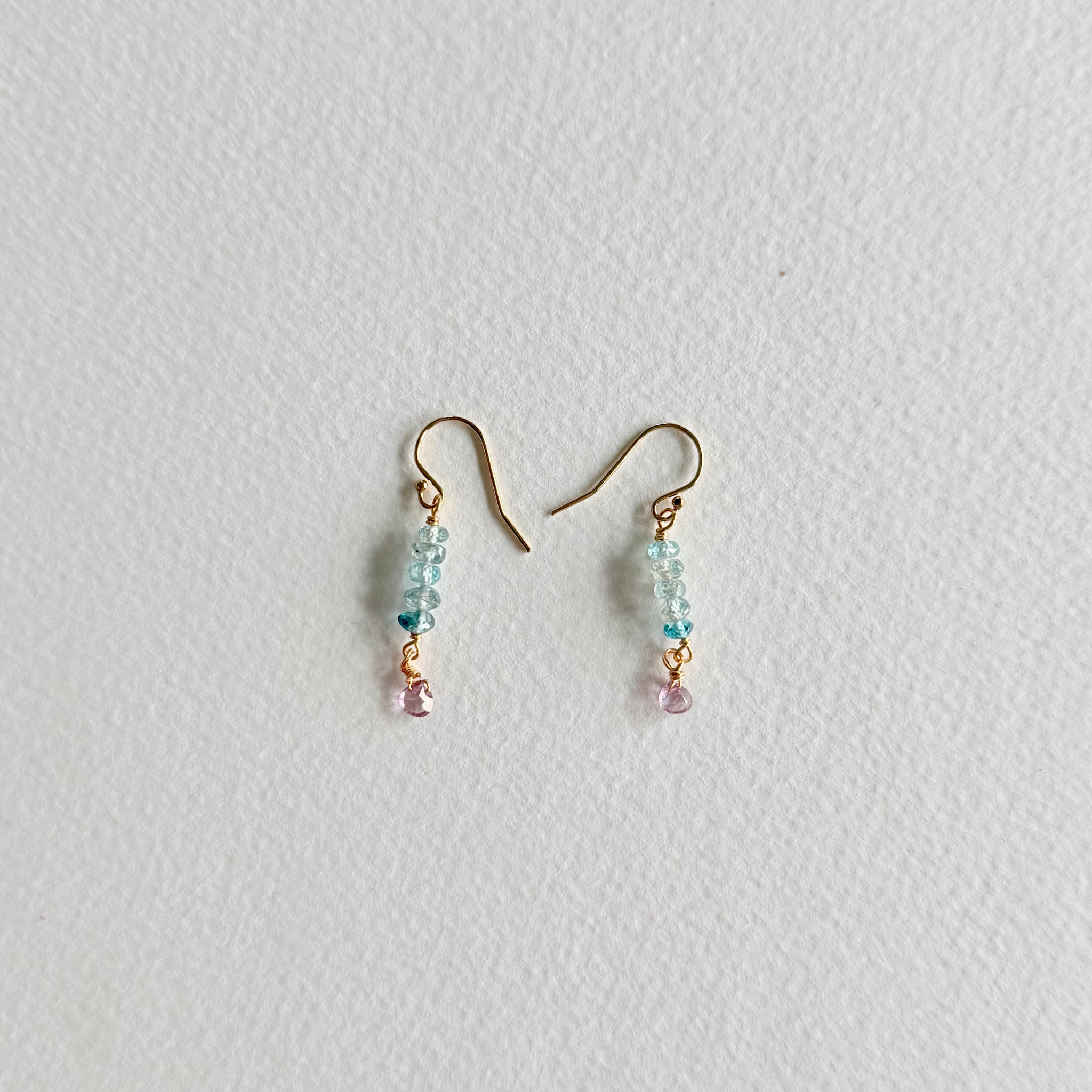 A pair of small dangle earrings with gold-tone hooks, light blue faceted beads, and tiny pink dangling accents, photographed on a plain light background.