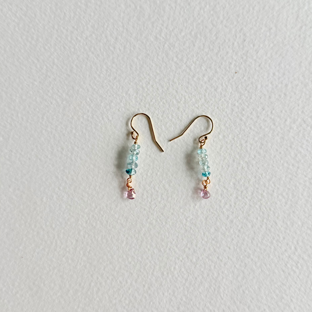 A pair of small dangle earrings with gold-tone fishhook wires, light blue faceted beads, a tiny dark blue bead, and a dangling light pink bead, shown on a textured off-white background.