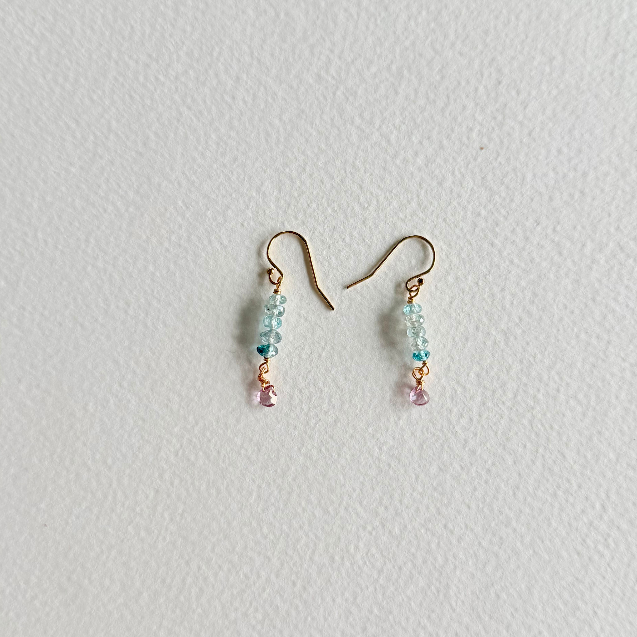 A pair of small dangle earrings with gold-tone fishhook wires, light blue faceted beads, a tiny dark blue bead, and a dangling light pink bead, shown on a textured off-white background.