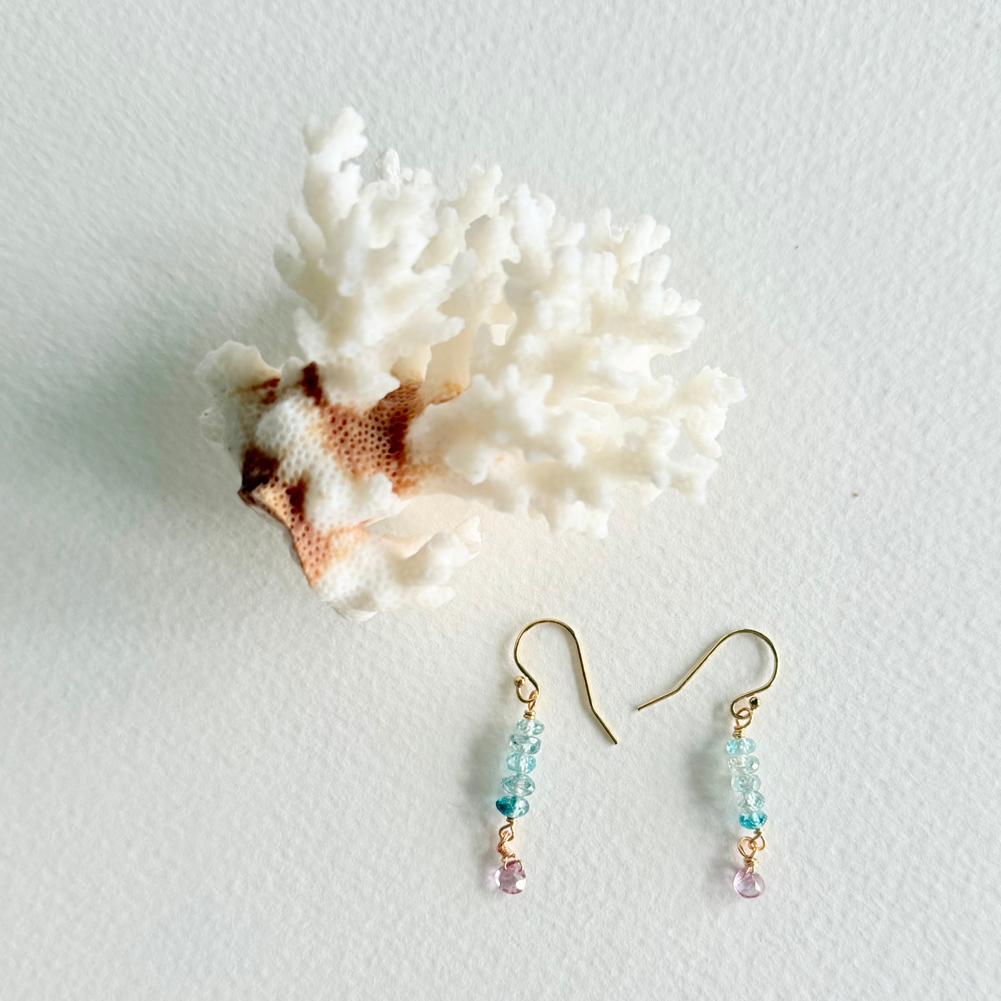 A pair of gold-tone dangle earrings with light blue and pink beads, displayed next to a white coral shell on a textured white background.