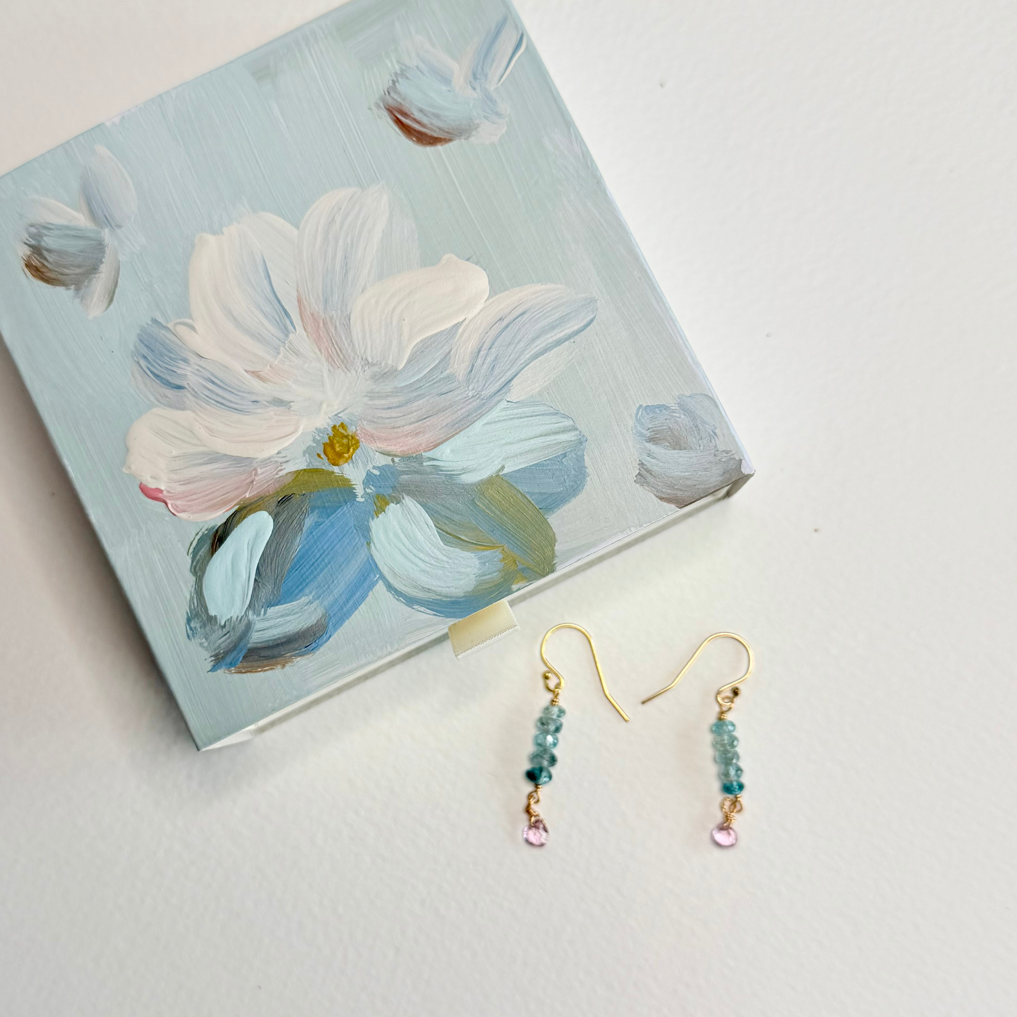 A pair of gold-tone dangle earrings with light blue beads and pink teardrop charms, displayed next to a small square canvas painting of a white and blue flower on a white background.