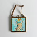 A square framed original art featuring an abstract watercolor painting of a dog in shades of turquoise, beige, and brown, suspended by a brown leather strap against a white wall.