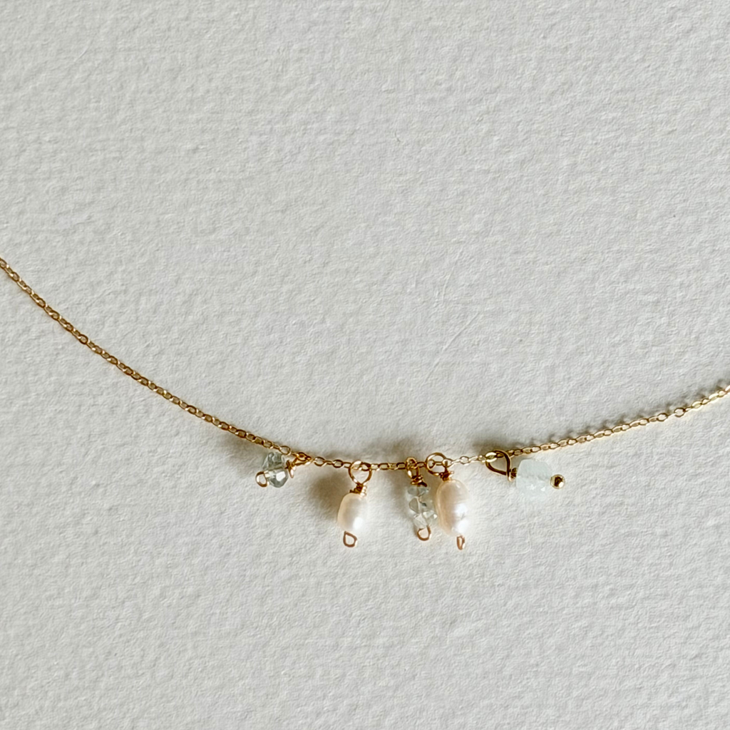A delicate gold chain necklace featuring three small pearl-like bead drops and tiny clear crystal charms, displayed on a textured off-white background.