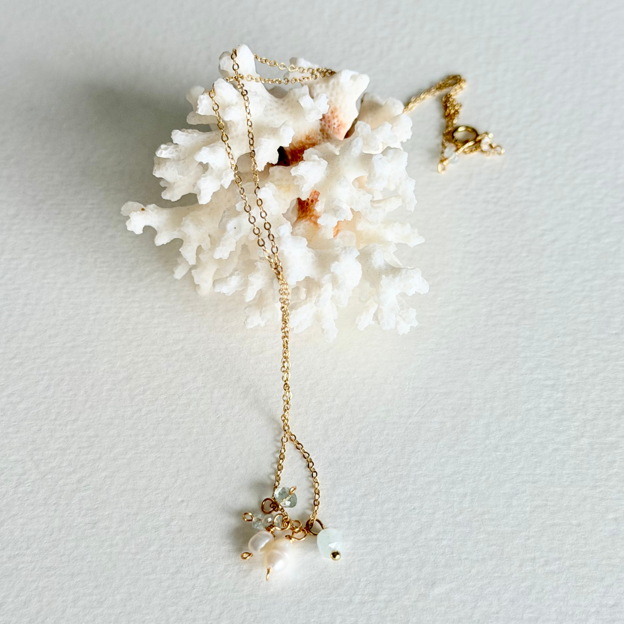 A delicate gold chain necklace draped over a white coral shell, featuring small pearl-like beads and a light blue bead accent at the pendant, set against a textured off-white background.