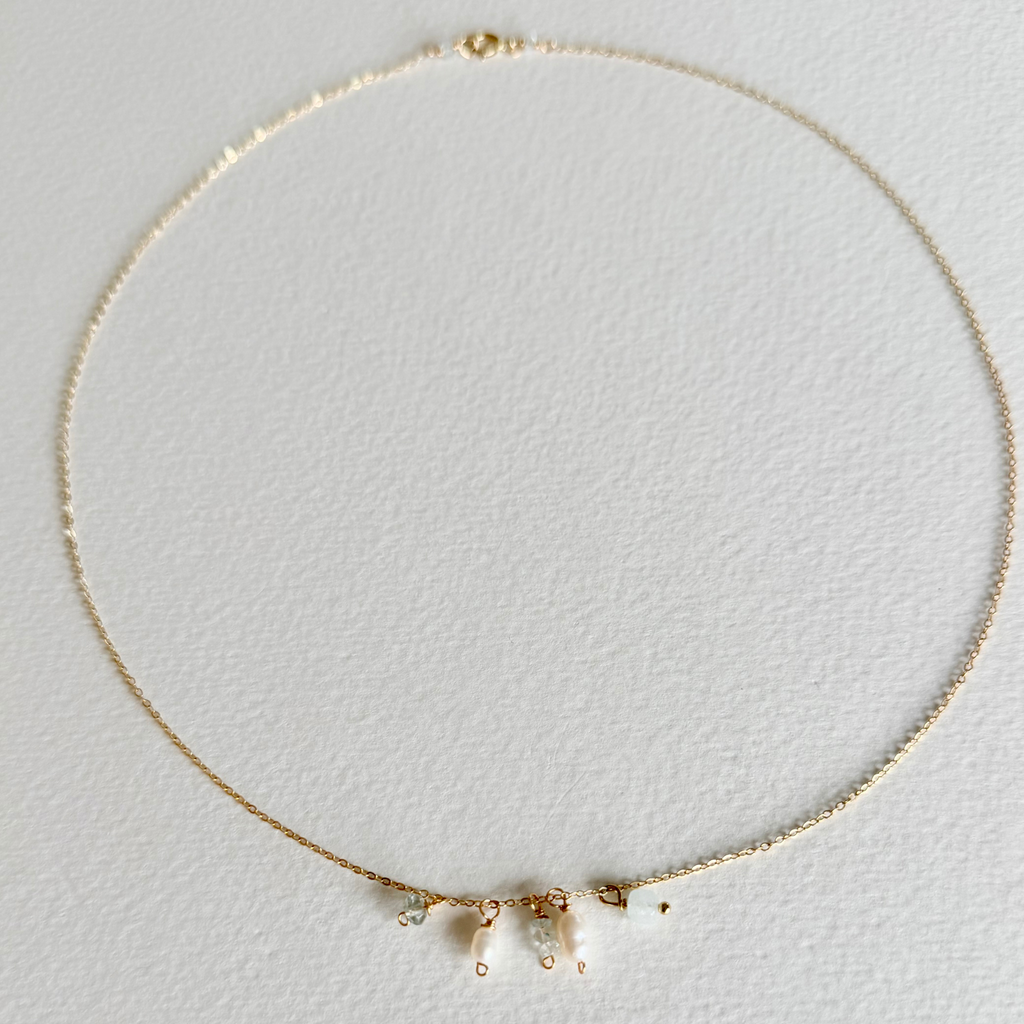 A delicate gold chain necklace with three small dangling charms, including a pearl and two tiny clear stones, displayed on a textured white background.