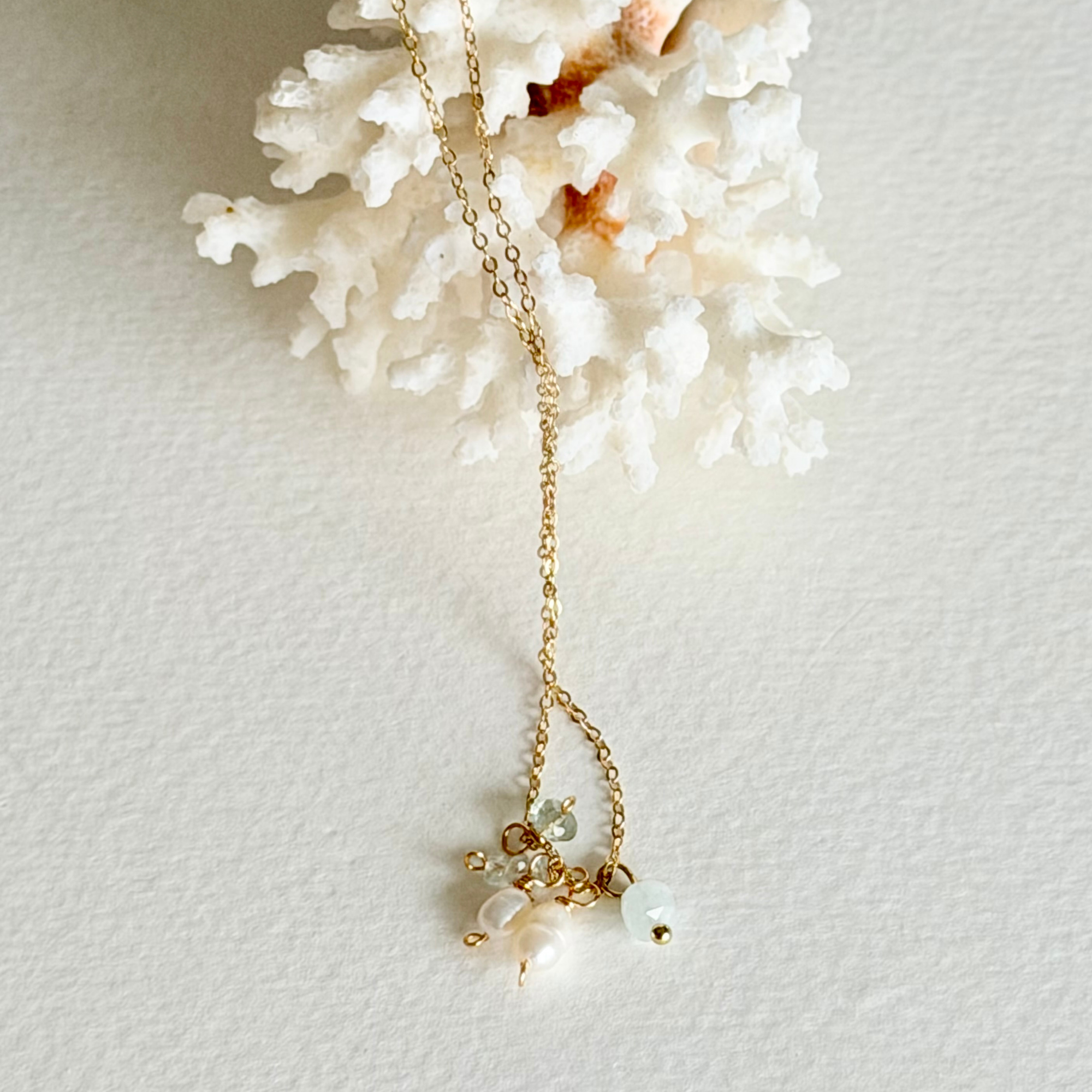 A delicate gold chain necklace with small pearl-like beads and a light green bead pendant, styled on a textured white surface next to a white coral fragment.