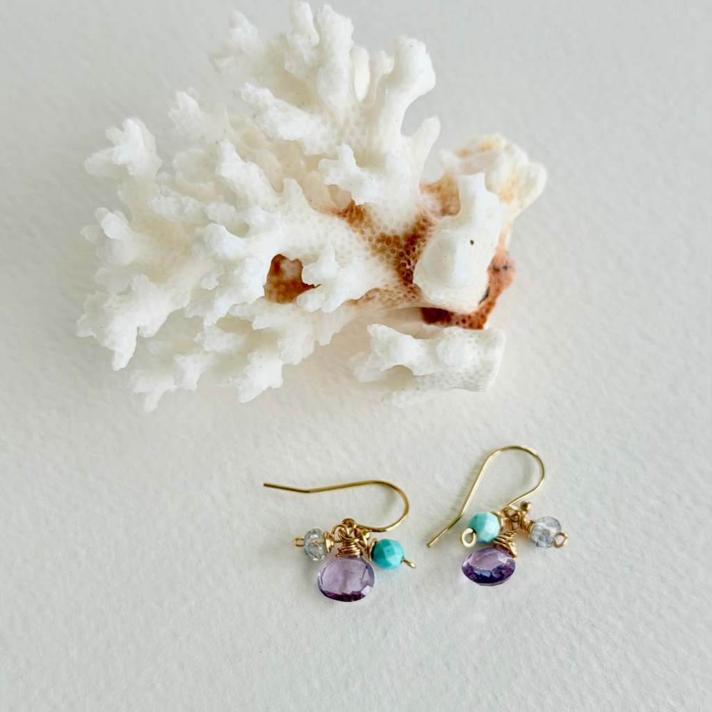 Pair of gold earrings with purple and turquoise beads on a white surface with coral.