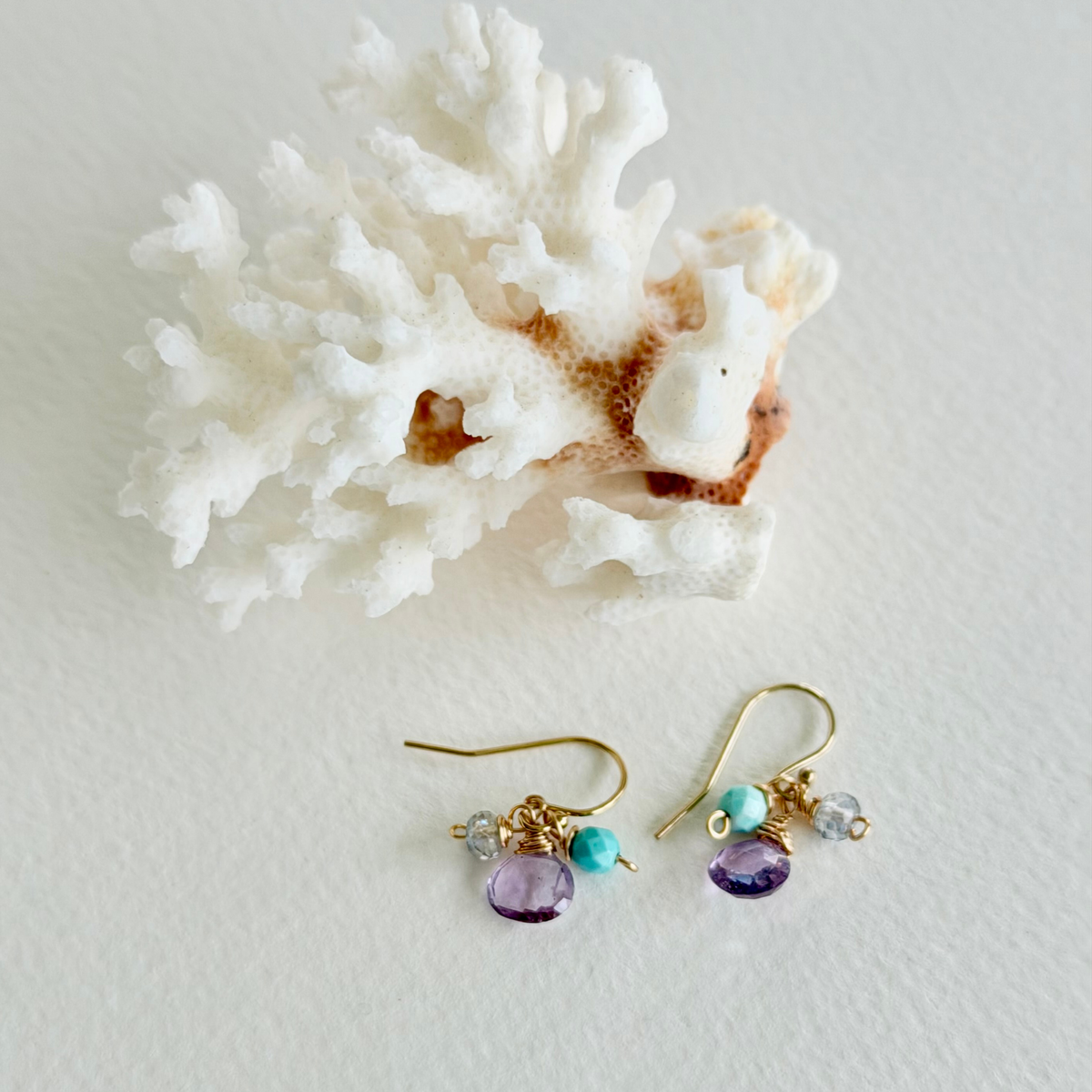 Pair of gold earrings with purple and turquoise beads on a white surface with coral.