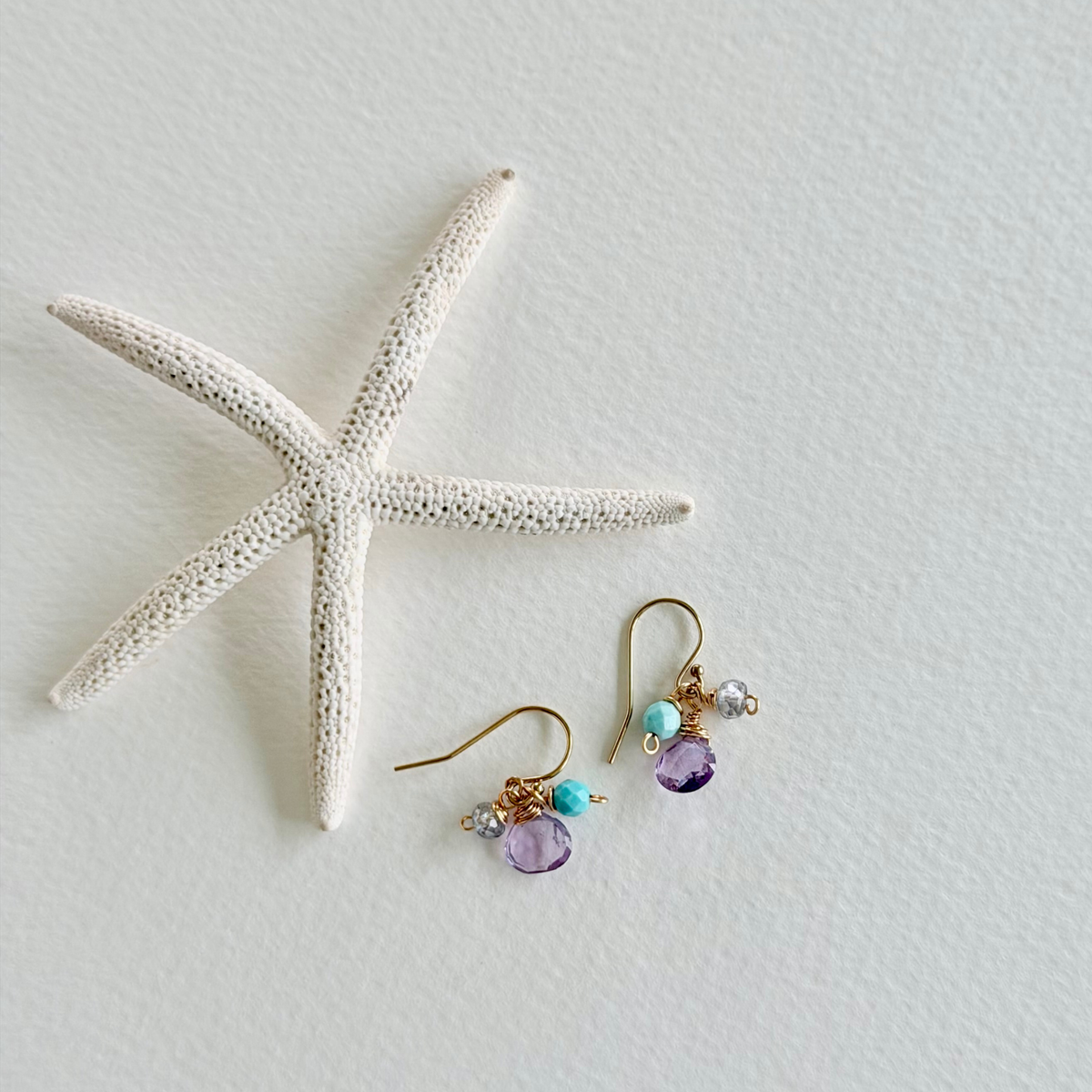 Pair of gold earrings with purple and blue beads on a textured white surface with a starfish.