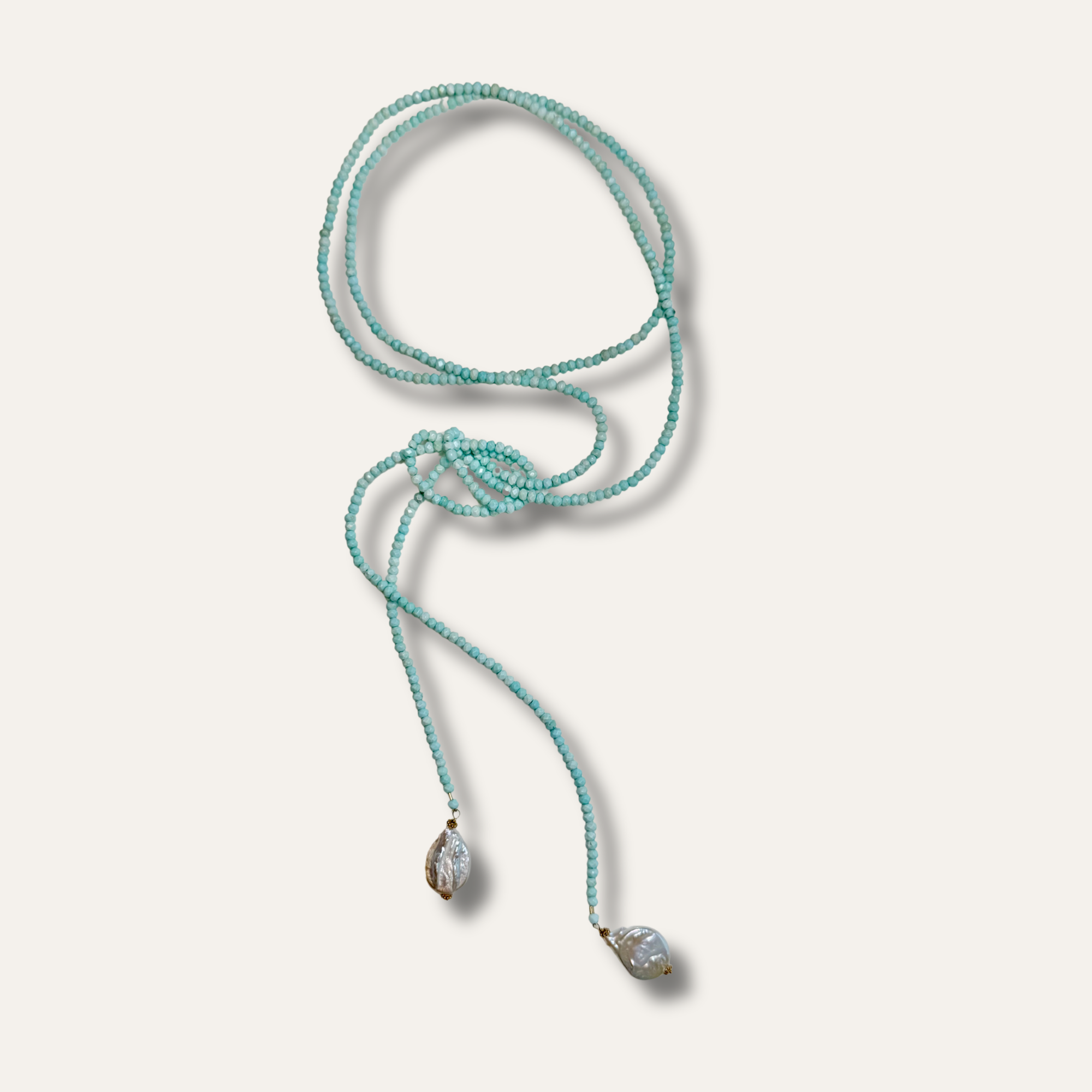 A turquoise beaded lariat necklace with two teardrop-shaped clear crystal pendants on a light beige background.