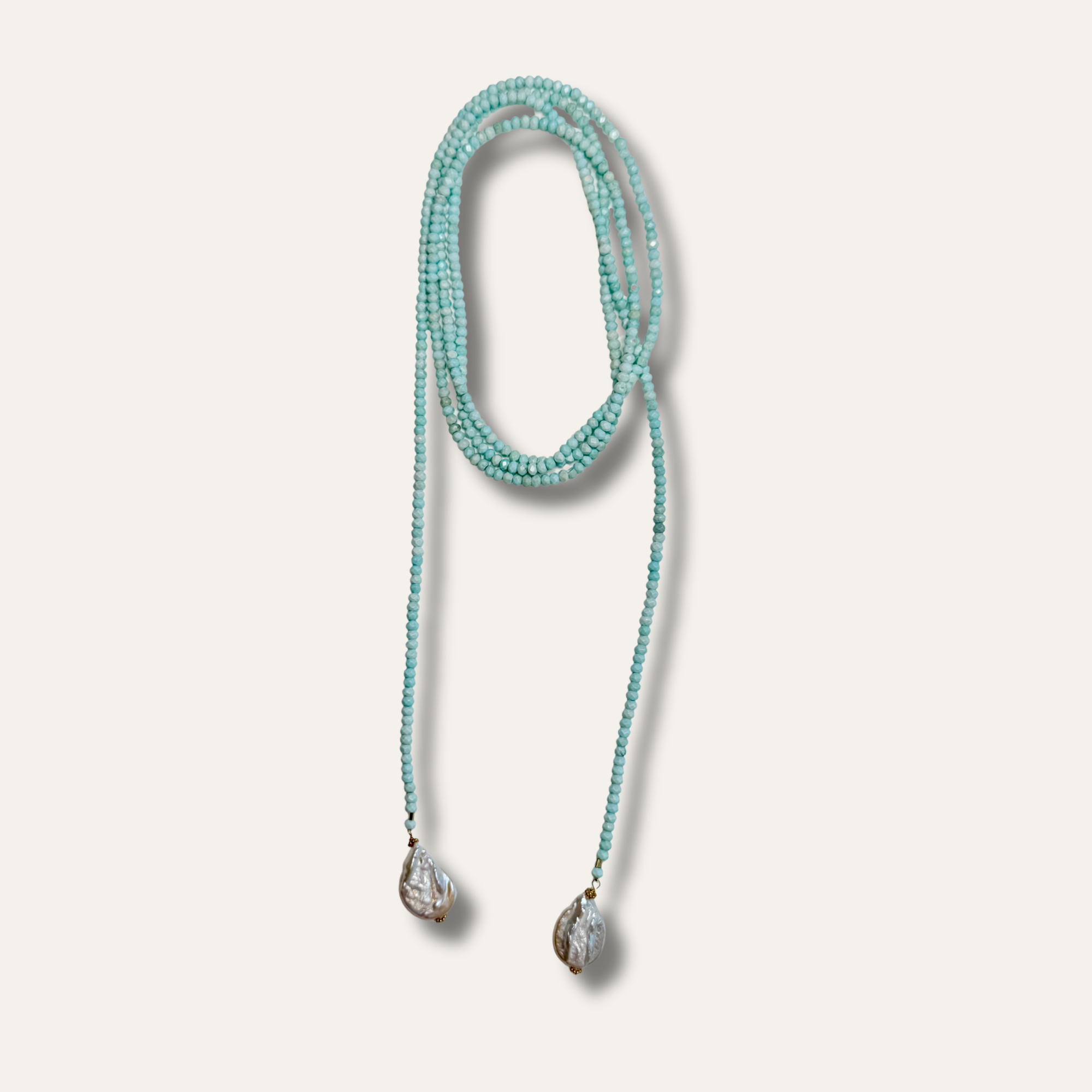 A long strand necklace made of small turquoise-blue beads, coiled in a loop with two dangling teardrop-shaped clear pendants on a light beige background.