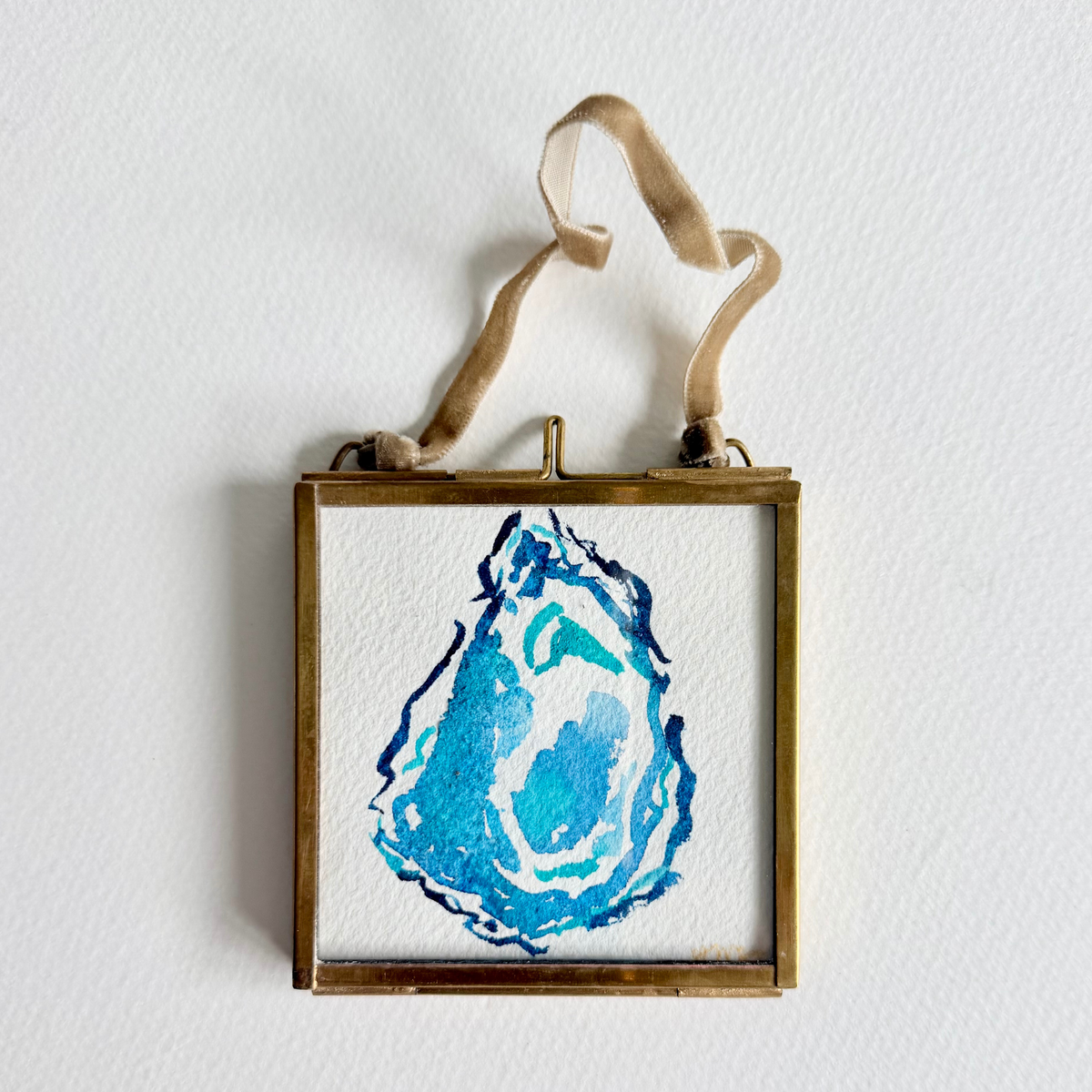A square framed original art featuring a blue and turquoise geode illustration on textured white paper, suspended by a beige ribbon from a brass frame against a white wall.