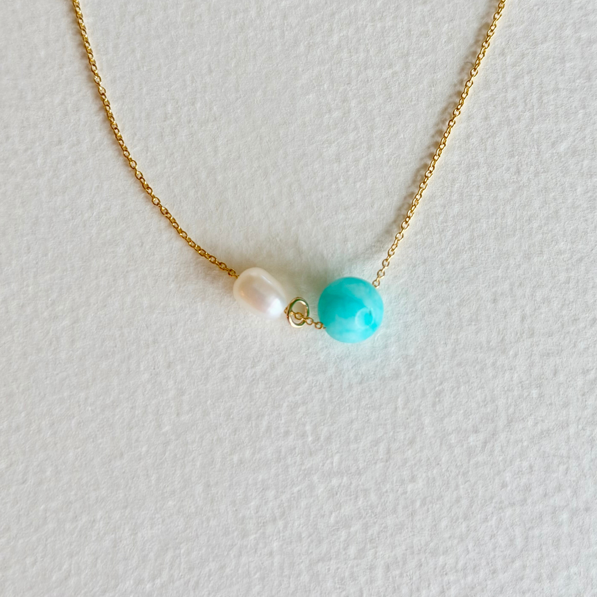 Necklace with a turquoise bead and pearl on a gold chain against a light gray background