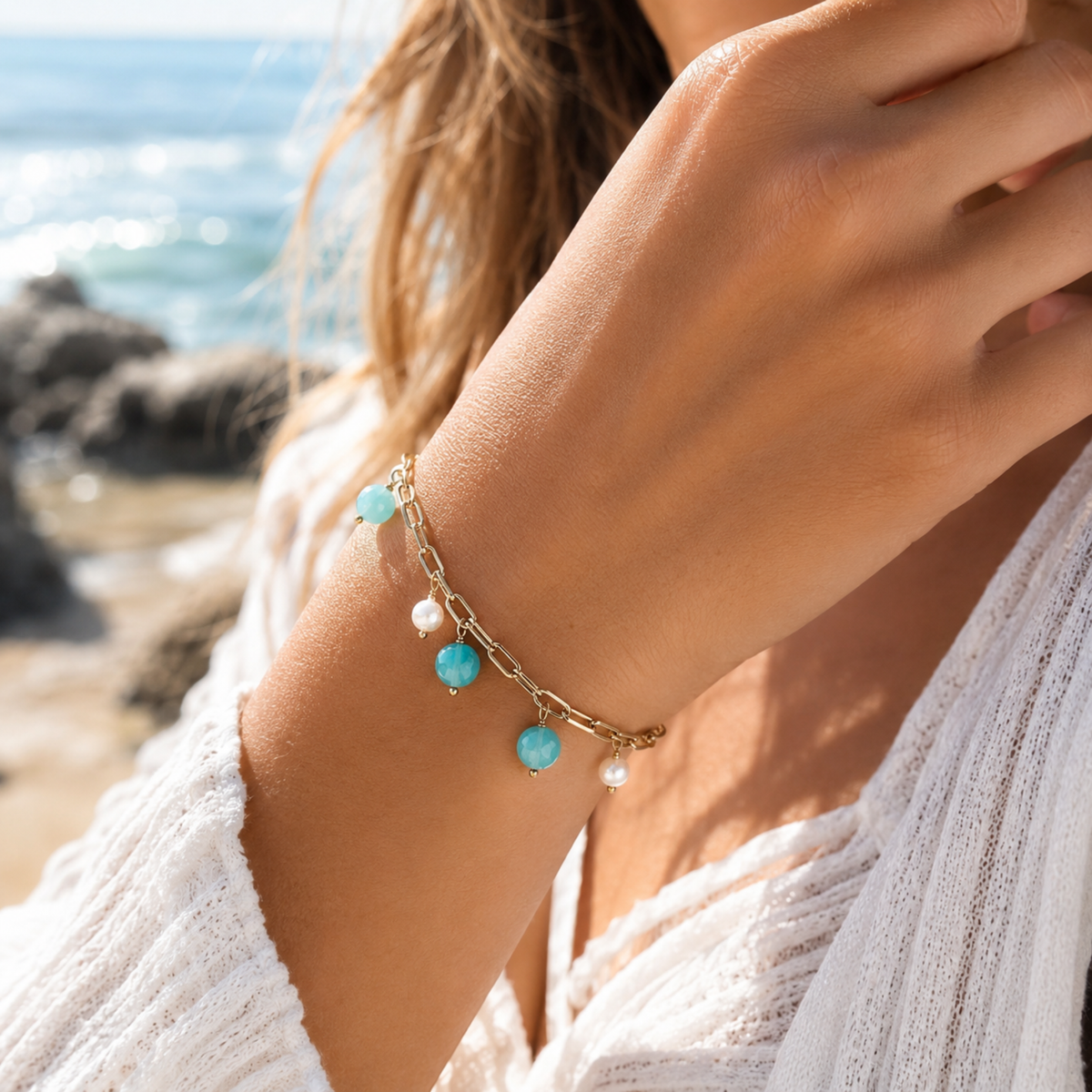 Close-up of a hand wearing a gold bracelet with amazonite beads and pearls against a beach background.