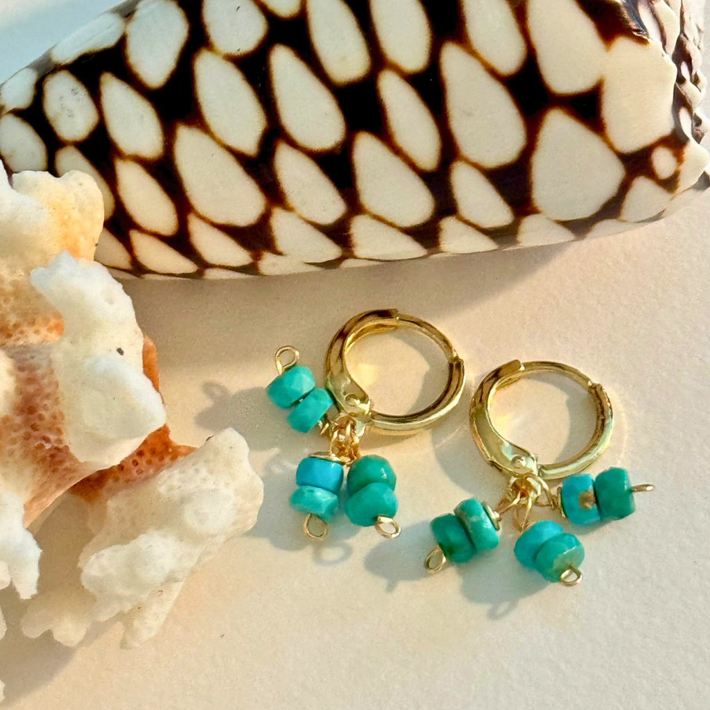 Up close view of a pair of earrings with a trio of genuine sleeping beauty turquoise on each hoop handcrafted with 14k gold-filled wire huggie clasp. Displayed between creamy white coral and brown and white spotted shell.