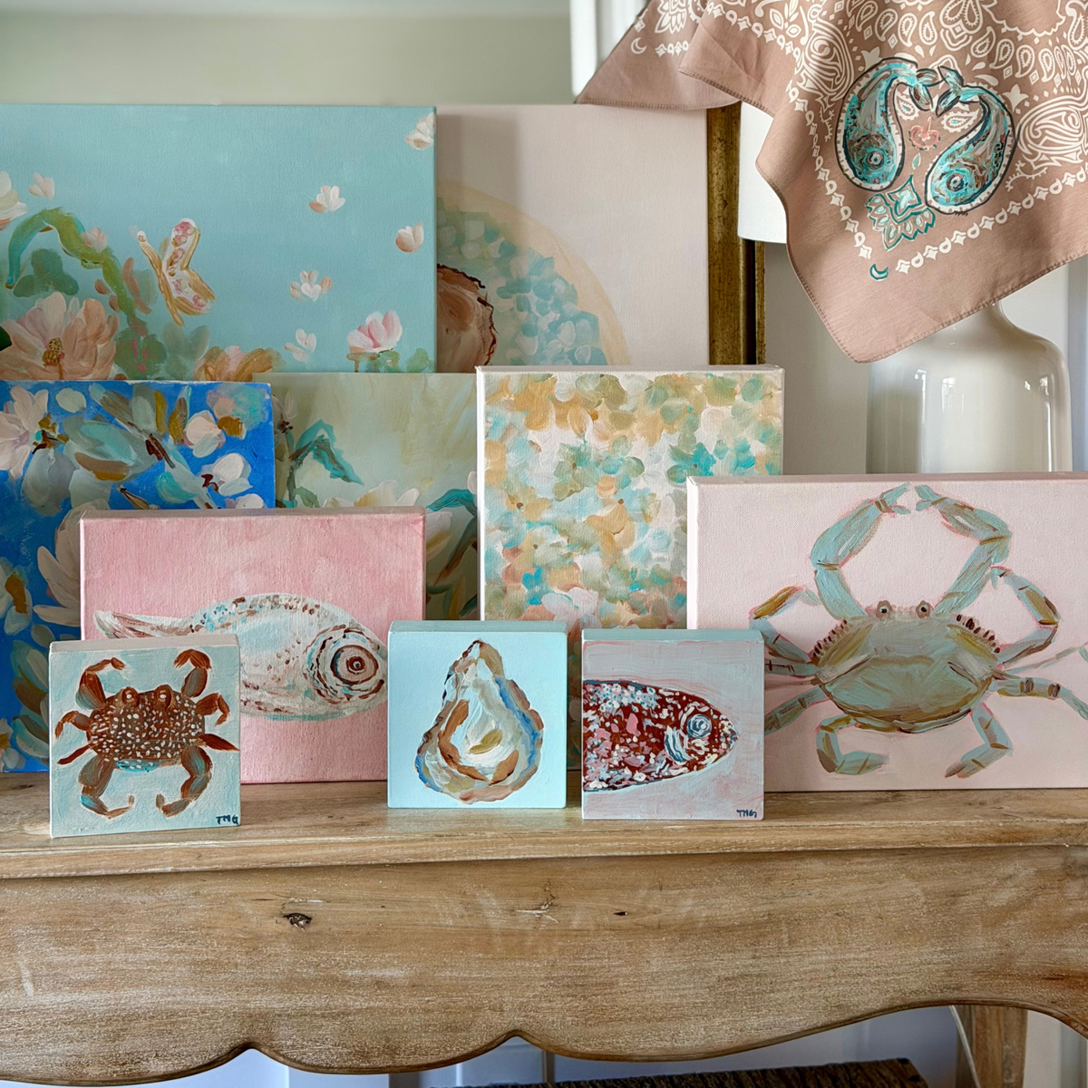 Collection of original coastal paintings by Tracy Marie Grogan, including blue crab, fish, oyster and floral artwork displayed with a hand-painted bandana.