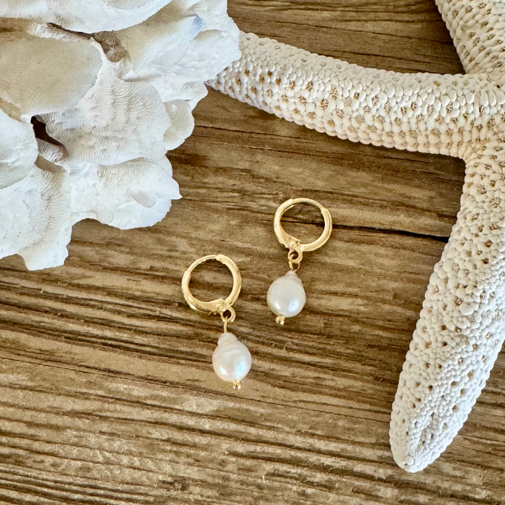 Pair of earrings with a single teardrop pearl on each hoop handcrafted with 14k gold-filled wire huggie clasp. Displayed between creamy white coral and starfish on a beachy wood grain surface.