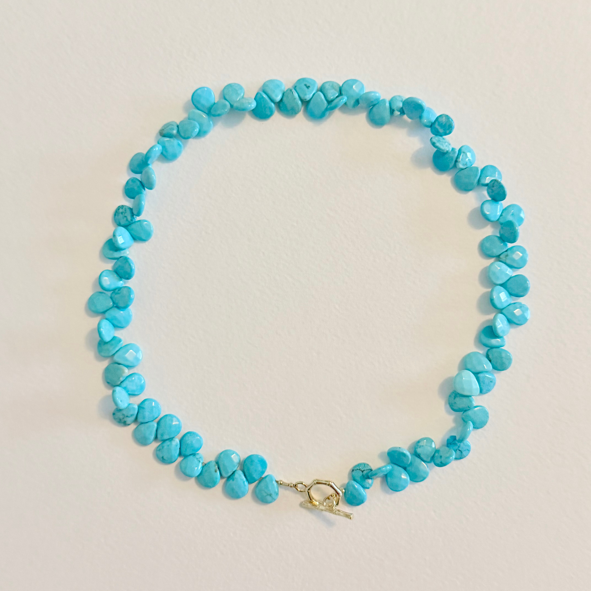 A turquoise beaded necklace with irregular, faceted beads and a small gold toggle clasp, laid flat on a light beige background.