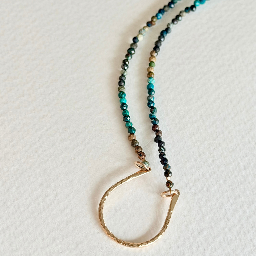 A beaded necklace with faceted beads in shades of teal, green, brown, and black, featuring a textured gold-toned clasp and a small gold-toned accent bead, laid on a textured off-white surface.