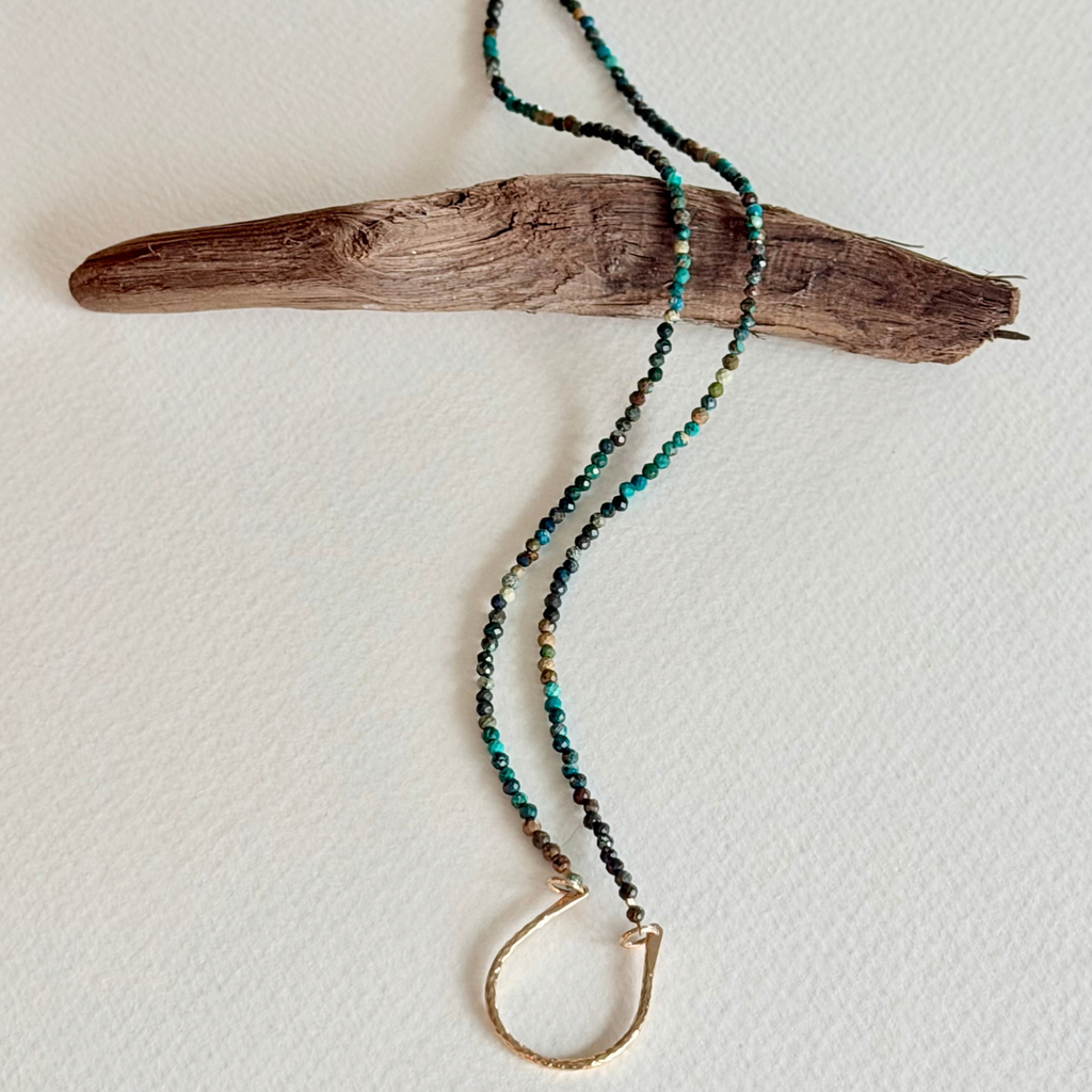 A beaded necklace with turquoise, black, and gold beads, featuring a gold-toned half-ring clasp, draped over a piece of driftwood on a textured off-white background.