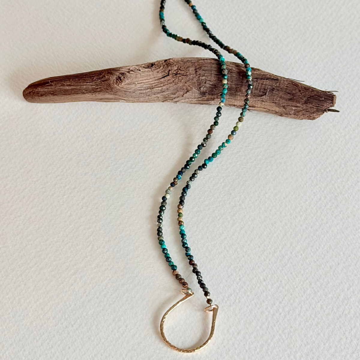 A beaded necklace with turquoise, black, and gold beads, featuring a gold-toned half-ring clasp, draped over a piece of driftwood on a textured off-white background.