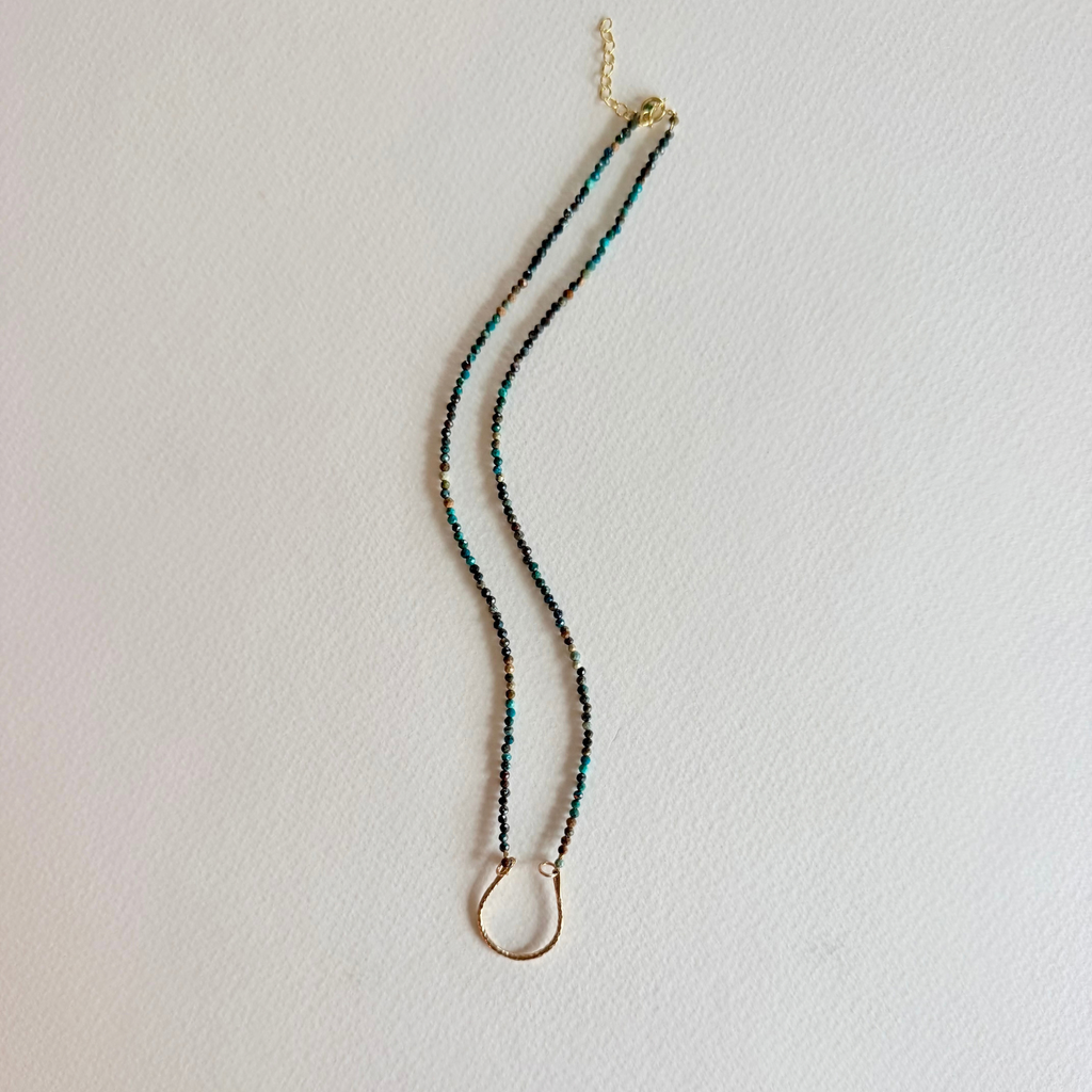 A delicate beaded necklace with small turquoise blue and gold beads, featuring a gold ring pendant and clasp, displayed against a plain white wall.