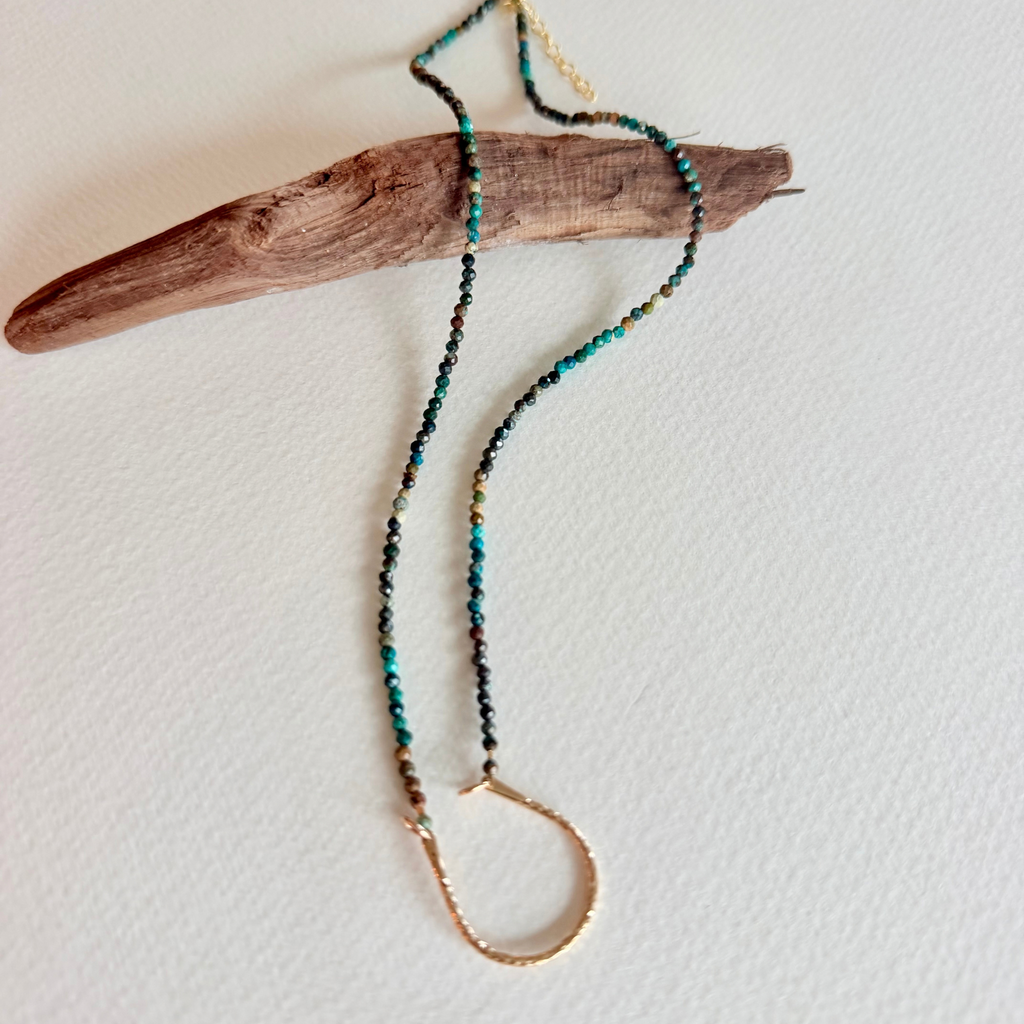 A beaded necklace with turquoise, black, and gold beads, featuring a textured gold-toned metal infinity loop pendant, draped over a piece of driftwood on a white background.