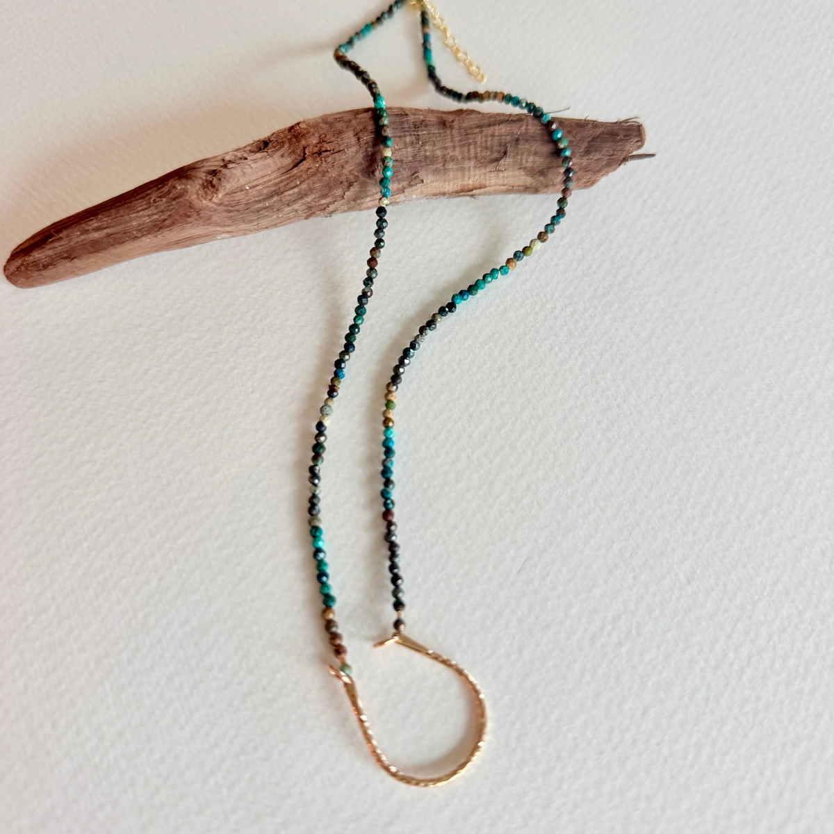 A beaded necklace with turquoise, black, and gold beads, featuring a textured gold-toned metal infinity loop pendant, draped over a piece of driftwood on a white background.