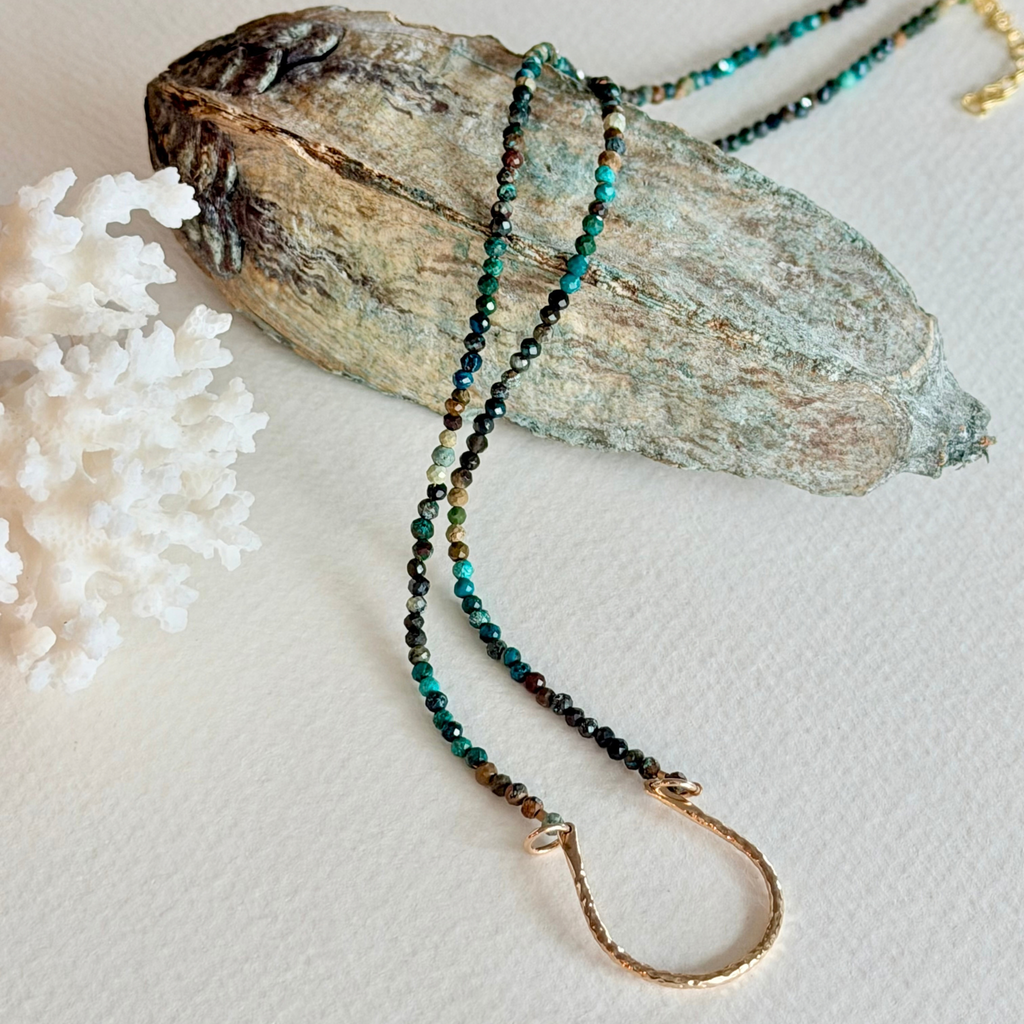 A beaded necklace with dark brown, black, and turquoise-green beads, featuring a gold-toned textured ring clasp, displayed on a piece of weathered wood with white coral.
