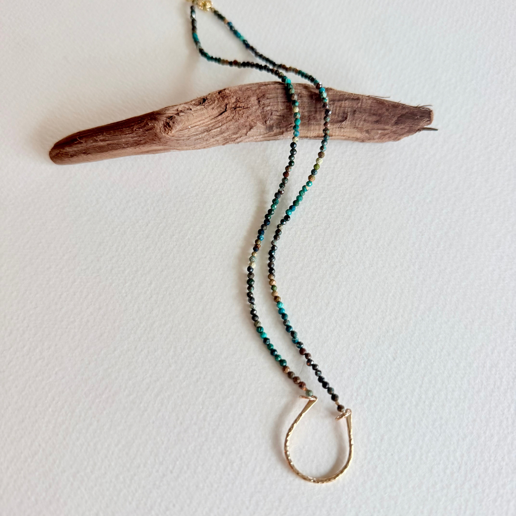 A beaded necklace with turquoise, black, and gold beads, featuring a gold-toned half-ring pendant, displayed on a piece of driftwood against a white background.