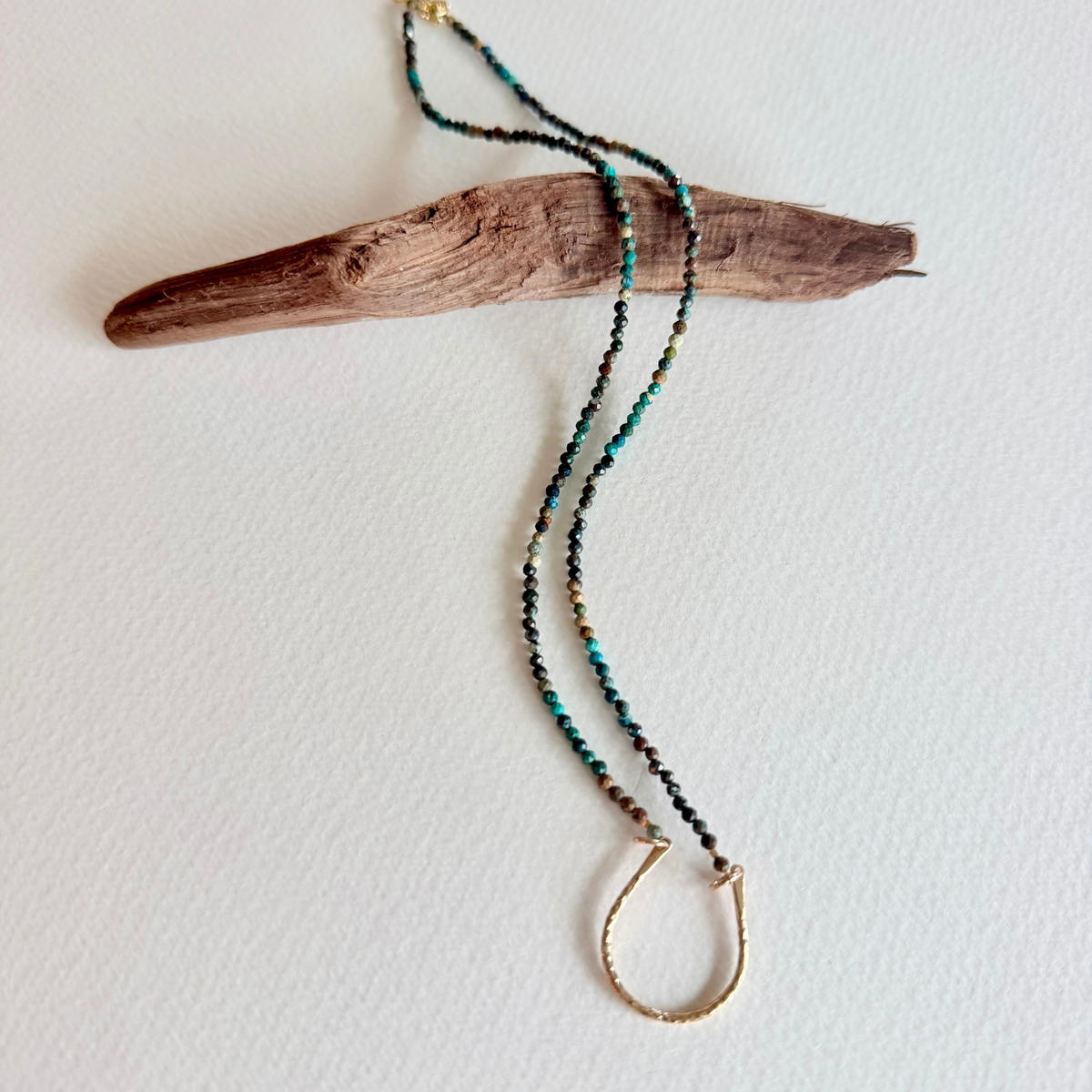 A beaded necklace with turquoise, black, and gold beads, featuring a gold-toned half-ring pendant, displayed on a piece of driftwood against a white background.
