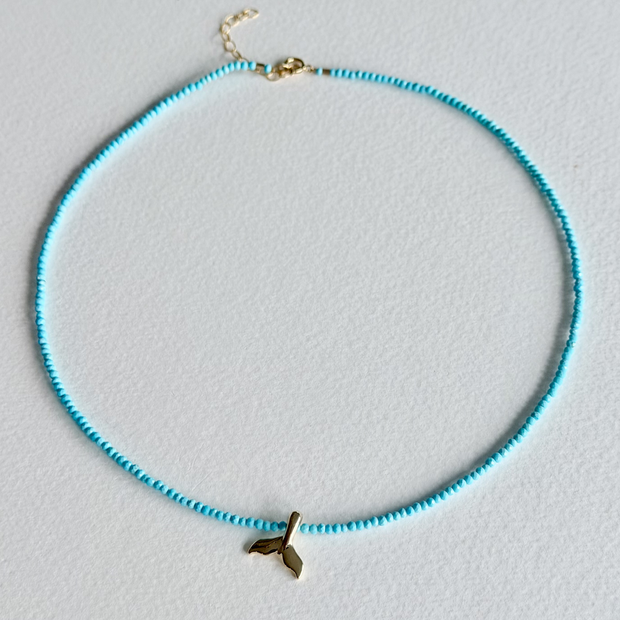 A delicate beaded necklace with light blue beads and a small gold whale tail charm, displayed on a clean white background.