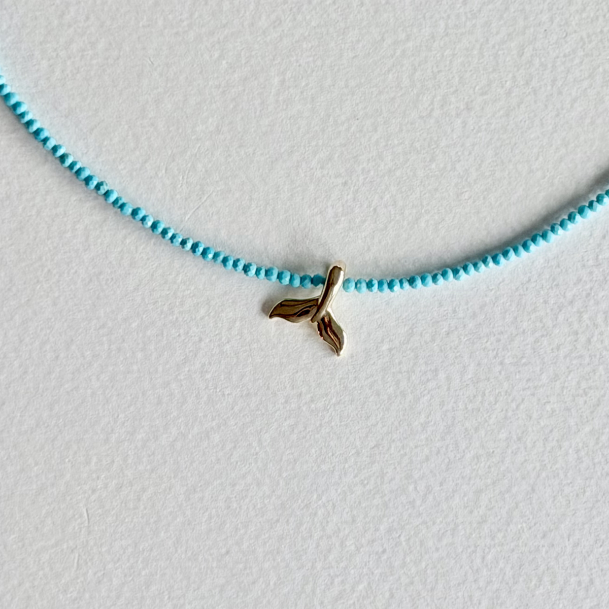 A delicate necklace featuring a small gold whale tail charm suspended on a thin, light blue beaded strand, displayed on a textured white surface.