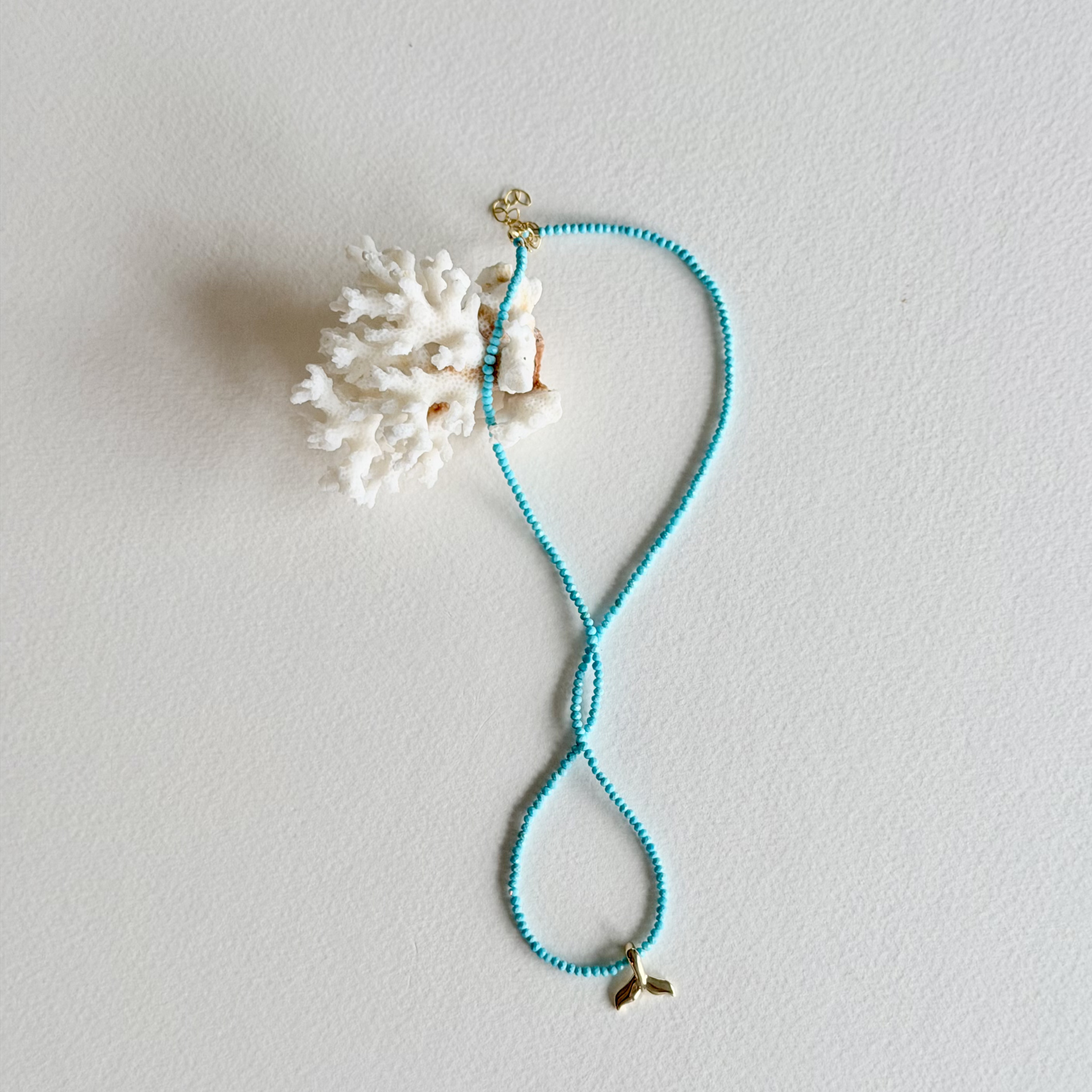A turquoise beaded necklace with a gold whale tail charm and a white coral shell pendant, displayed on a textured off-white background.