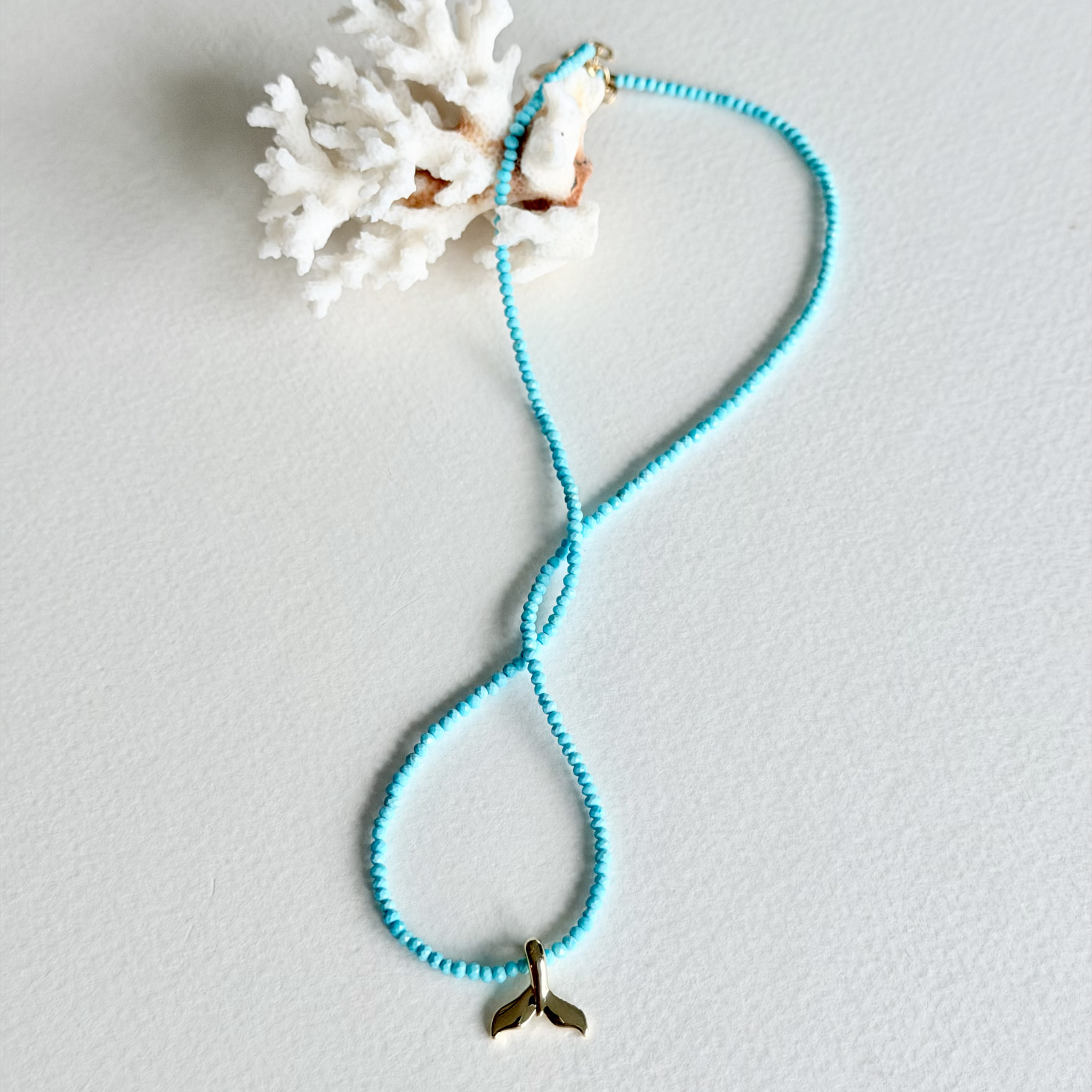 A turquoise blue beaded necklace with a small gold mermaid tail charm, displayed on a textured white surface next to a white coral shell.