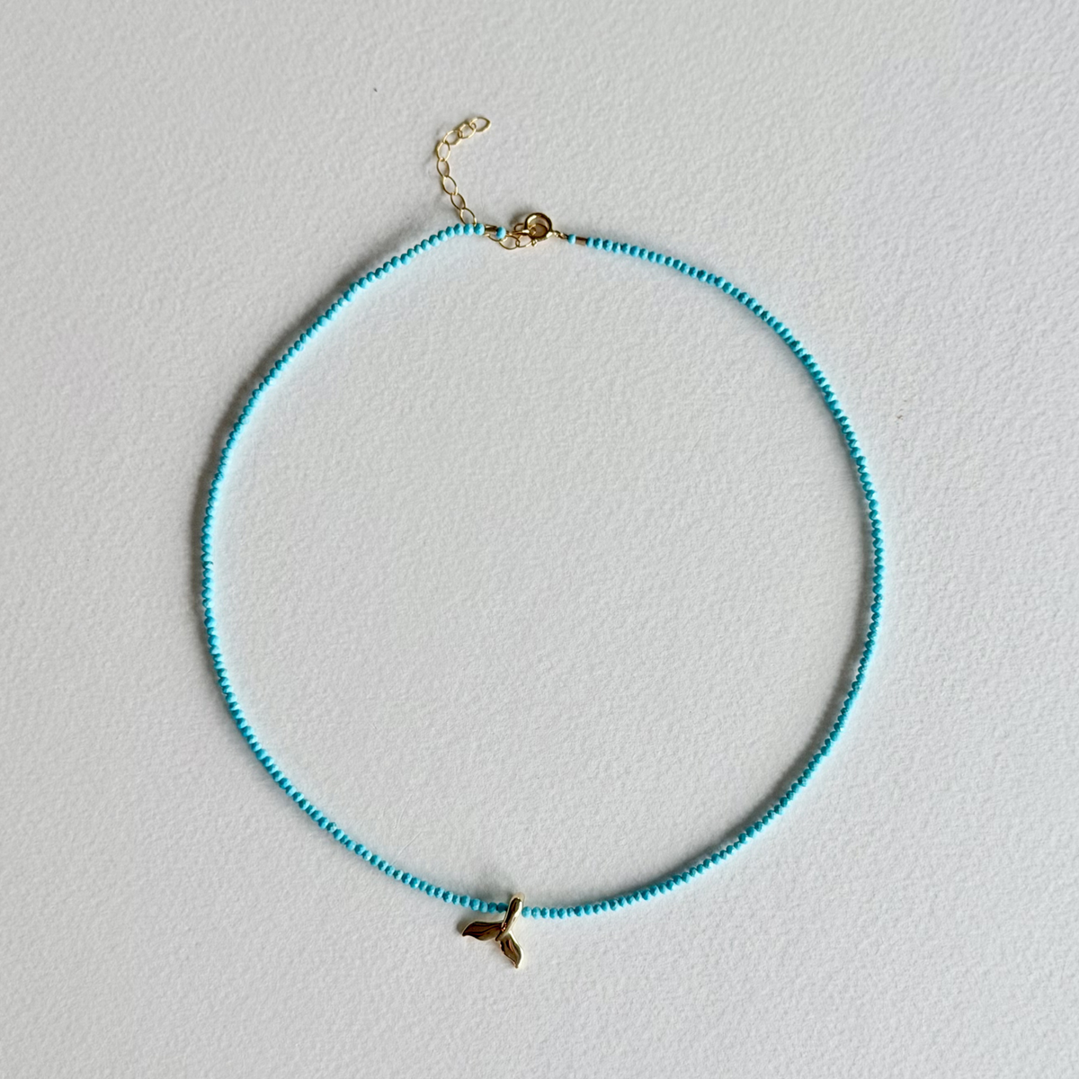 A delicate beaded necklace with small turquoise-blue beads and a gold mermaid tail charm, displayed on a textured white background.