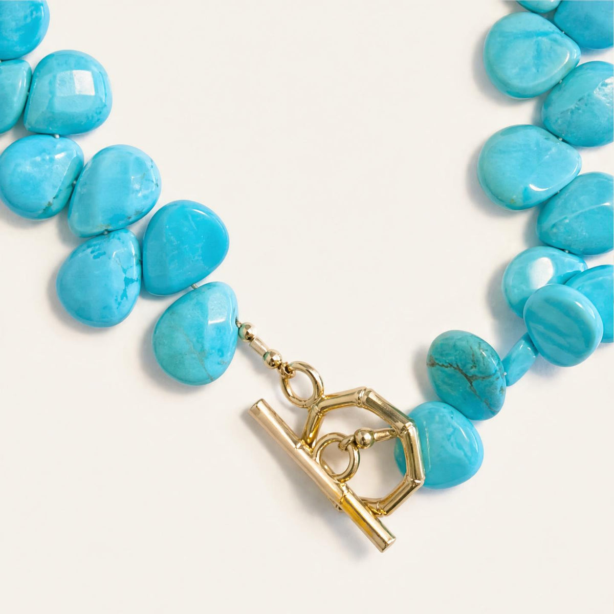 A close-up of a turquoise stone bead necklace with a gold toggle clasp on a light cream background.