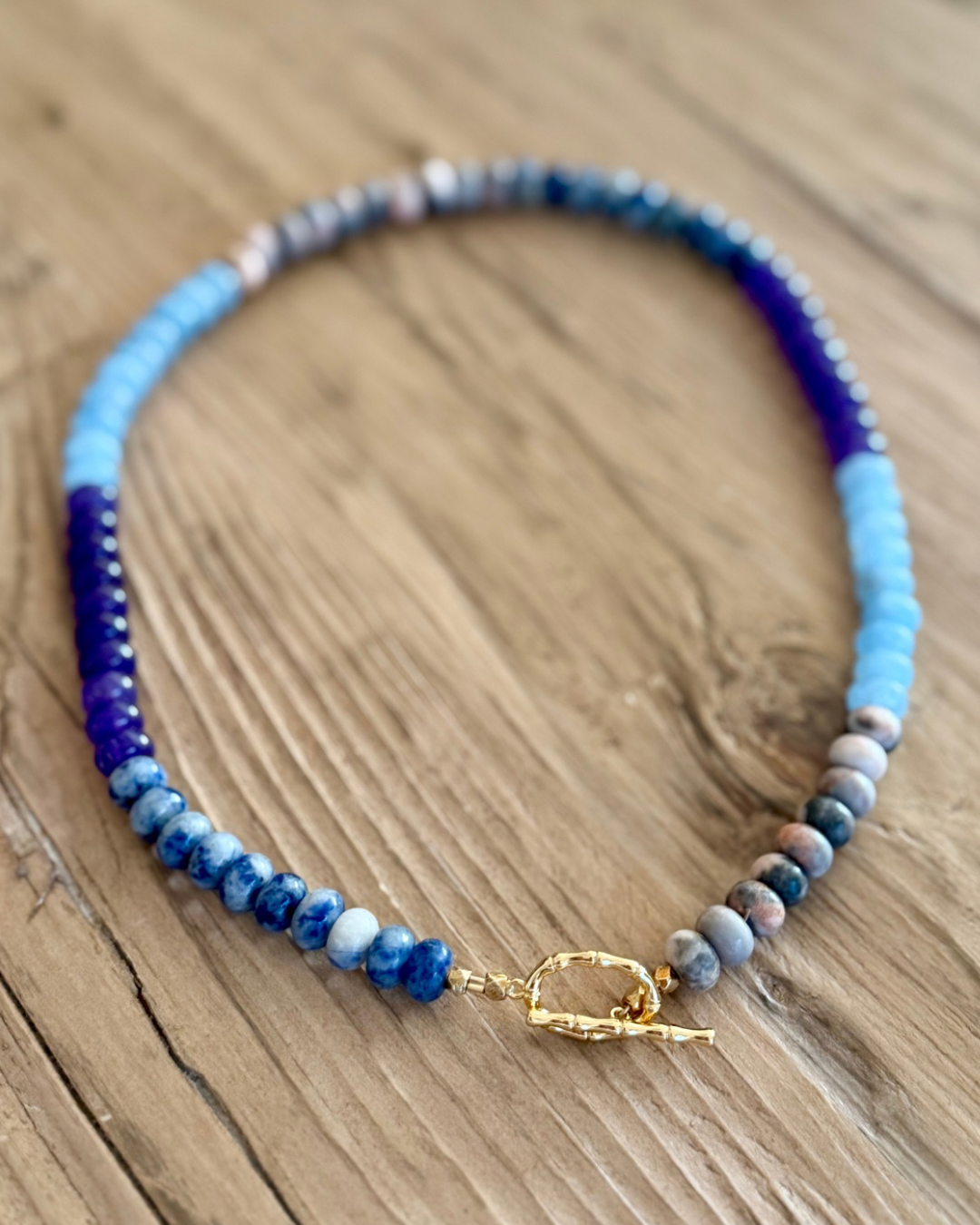 Violet Hour handmade 16-inch gemstone necklace featuring violet and blue jade, Blue Spot Jasper, Pink Zebra Jasper and gold toggle clasp on rustic wood.