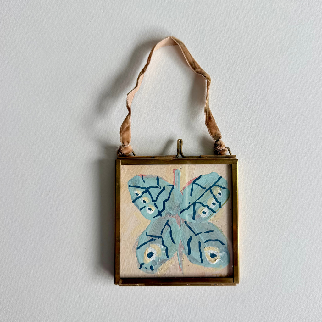 A square framed wall hanging with a light blue butterfly painted on a beige background, suspended by a tan ribbon against a white wall.