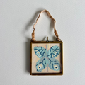 A square framed wall hanging with a light blue butterfly painted on a beige background, suspended by a tan ribbon against a white wall.