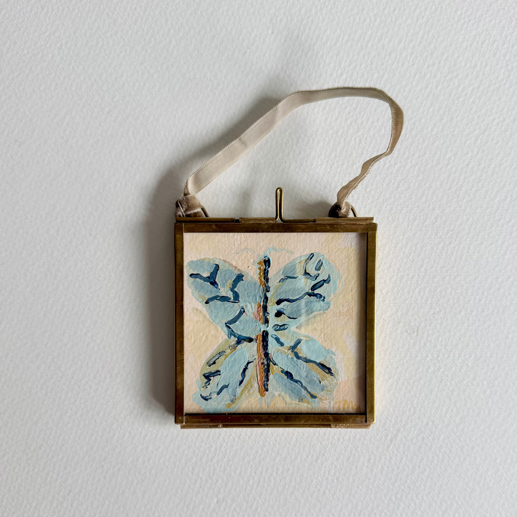 A square framed hanging frame with a small butterfly painting in light blue and navy on a cream background, suspended by a beige ribbon against a white wall.