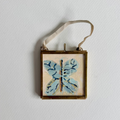 A square framed hanging frame with a small butterfly painting in light blue and navy on a cream background, suspended by a beige ribbon against a white wall.