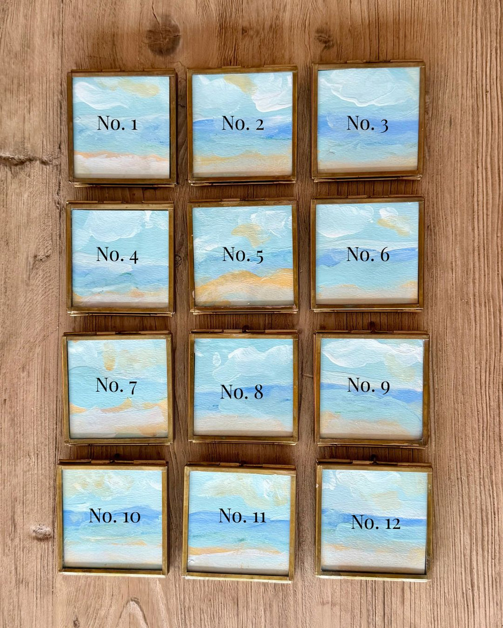 Twelve small square framed cards arranged in a 3-by-4 grid on a wooden surface. Each card has a light blue and beige painted ocean scene with black text reading 'No.