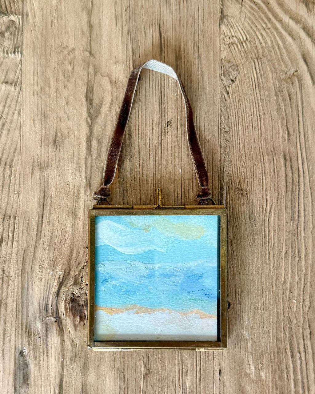 A small framed beach landscape painting with a brown leather hanging strap, displayed on a rustic wooden background.