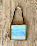 A small framed beach landscape painting with a brown leather hanging strap, displayed on a rustic wooden background.